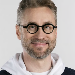 Silmälasipäinen mies katsoo hymyillen kameraan. Päällään hänellä on huppari.