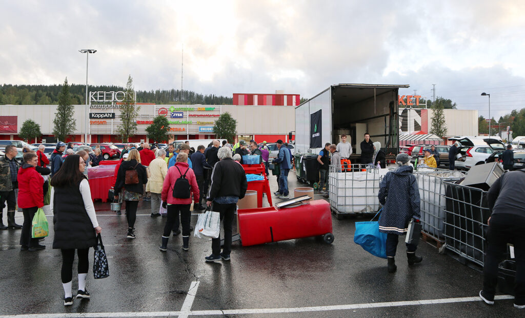 Kuvassa on jonkinlainen rekka ja jäteastioita sekä paljon ihmisiä. Taustalla näkyy iso rakennus, jossa lukee Keljon kauppakeskus.