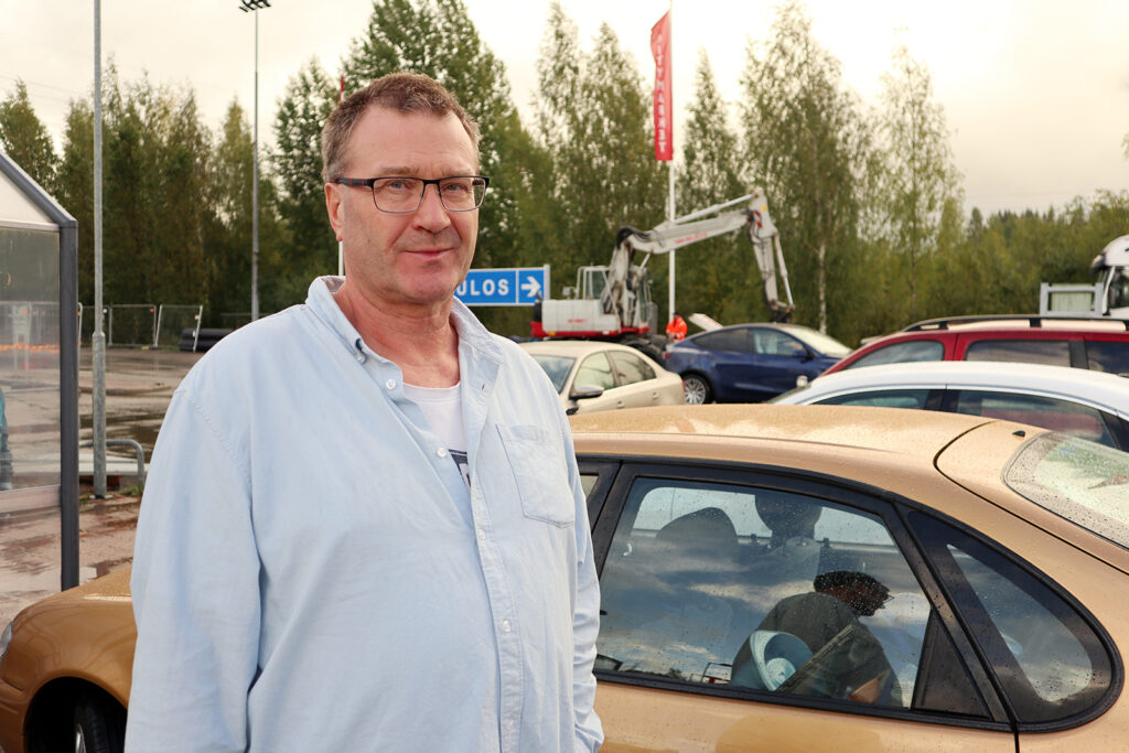 Mies kauluspaidassa katsoo kameraan. Takana näkyy autoja.