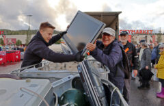 Lippalakkipäinen mies nostaa nuoremmalle miehelle isoa televisiota. Häntä naurattaa. Takana näkyy myös muita ihmisiä.