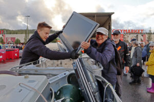 Lippalakkipäinen mies nostaa nuoremmalle miehelle isoa televisiota. Häntä naurattaa. Takana näkyy myös muita ihmisiä.