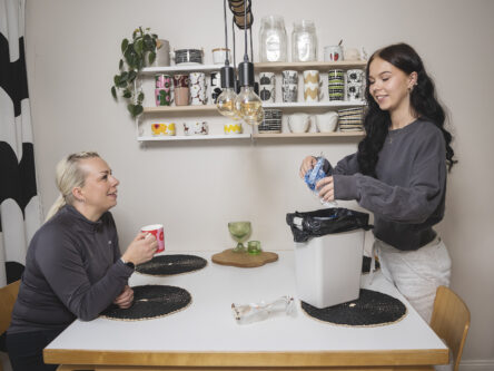 Vaaleahiuksinen nainen istuu keittiön pöydän ääressä ja juo kahvia Marimekko-mukista. Nuorempi ruskeahiuksinen tyttö laittaa pöydällä olevaan roskikeen muoviroskaa.
