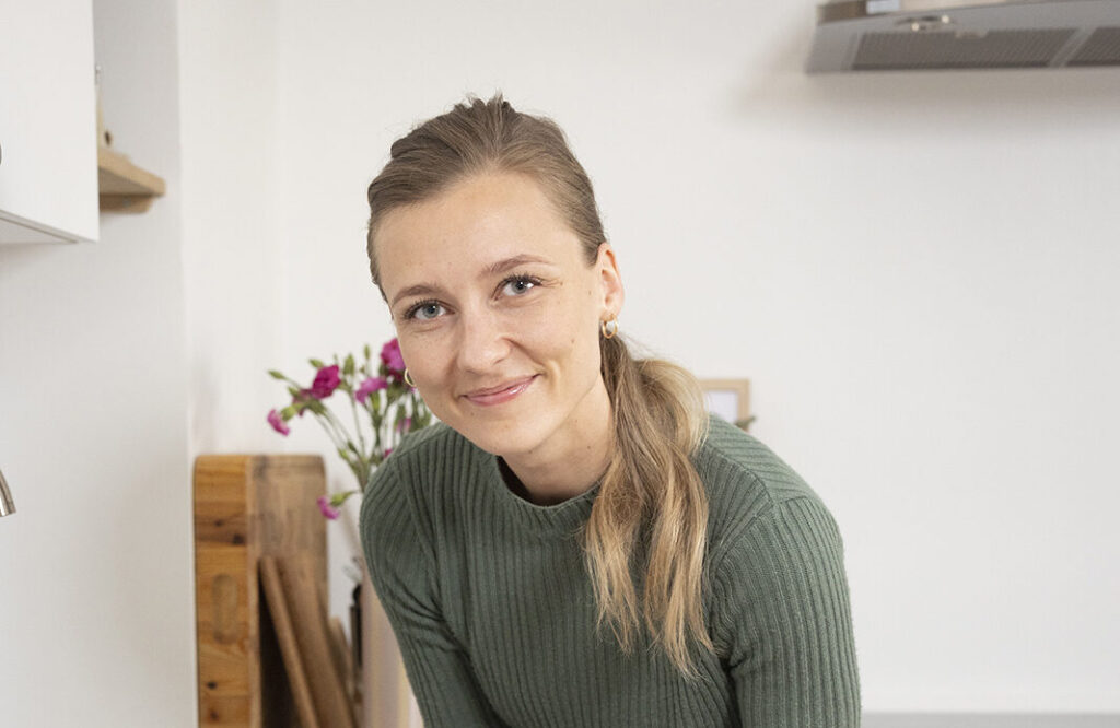 Nuori nainen on laittamassa roskikseen tyhjää muovipakkausta. Takana näkyy keittiötä ja pöydällä kukkia.