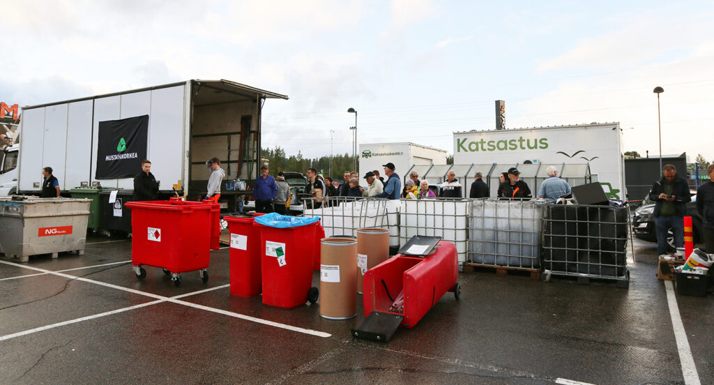 Vasemmassa reunassa on auto, jossa lukee Mustankorkea. Valkoisten jäteastioiden takana on rivissä ihmisiä odottamassa.