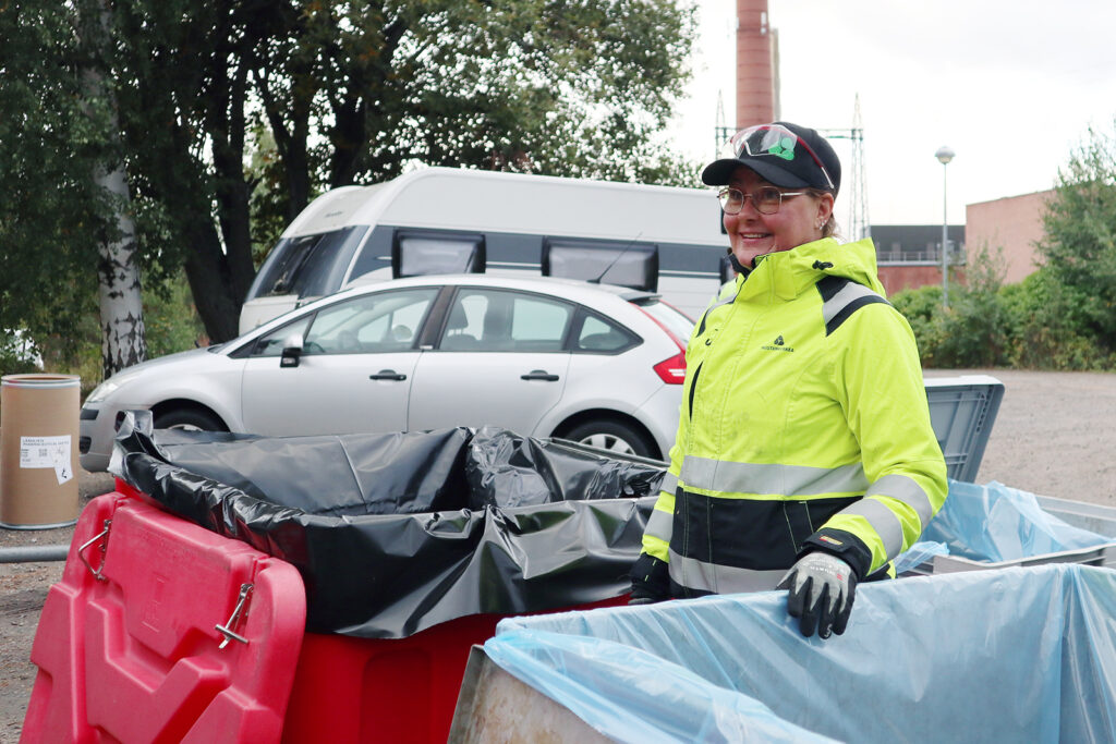 Huomiotakki päällä oleva nainen seisoo jäteastian vieressä ja hymyilee. Takana näkyy kaksi autoa.
