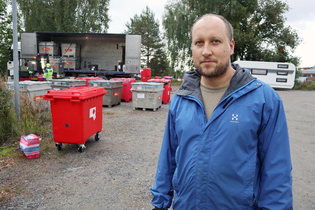 Sinitakkinen mies katsoo kameraan. Takana näkyy punaisia jäteastioita.