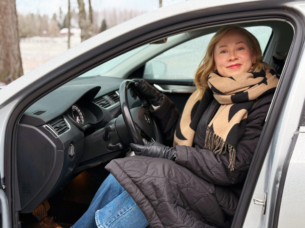 Vaaleahiuksinen nainen istuu autossa ratin takana ja hymyilee.