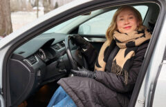 Vaaleahiuksinen nainen istuu autossa ratin takana ja hymyilee.