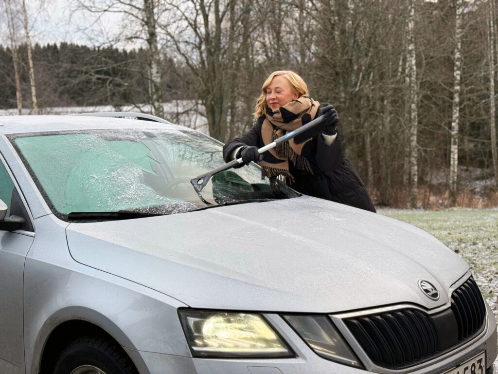 Vaaleahiuksinen nainen puhdistaa auton etuikkunaa jäästä. Maa on hieman kuurassa, joten on pientä pakkasta.