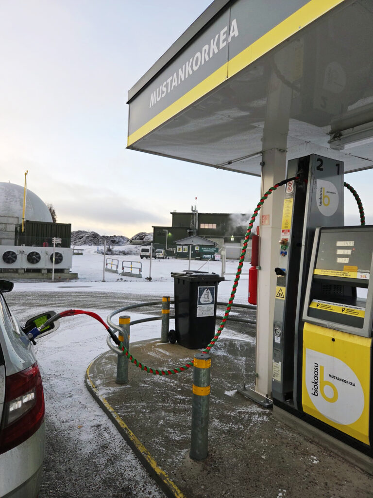 Kuvassa on Mustankorkean tankkausasema. Vieressä näkyy auto, jossa on tankkauspistooli ja takana vihreää rakennusta ja muuta tekniikkaa.