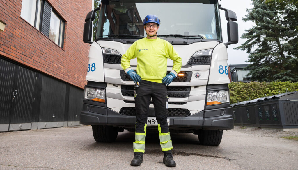 Mies pitää käsiä lanteillaan ja seisoo jäteauton edessä. Sivulla näkyy punatiilistä rakennusta, toisella puolella puita.