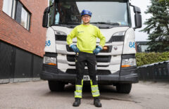 Mies pitää käsiä lanteillaan ja seisoo jäteauton edessä. Sivulla näkyy punatiilistä rakennusta, toisella puolella puita.