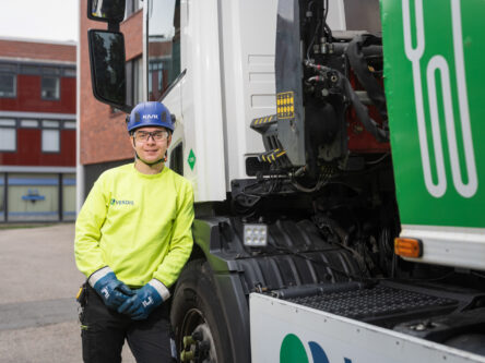 Nuori mies nojailee autoon ja hymyilee. Takana näkyy punaisia rakennuksia. Miehellä on päässään sininen kypärä, suojalasit ja keltainen huomiopaita.
