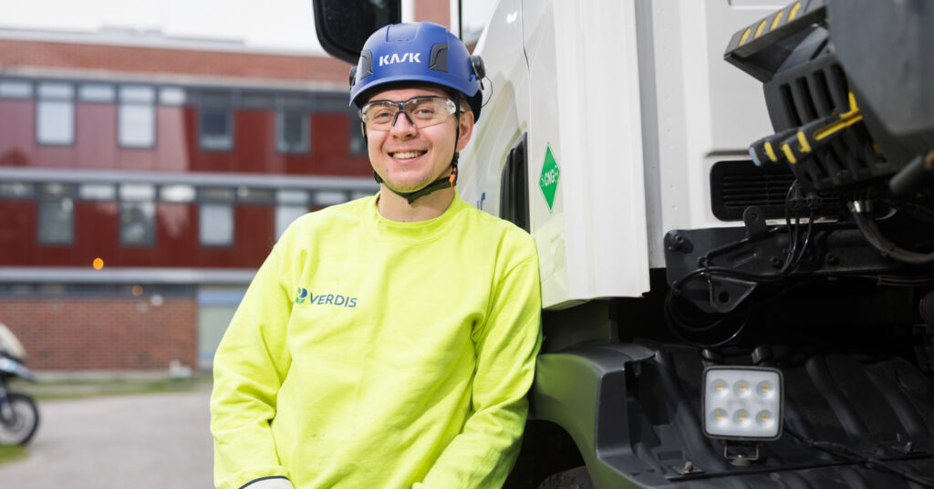 Nuori mies nojailee autoon ja nauraa. Takana näkyy punaisia rakennuksia. Miehellä on päässään sininen kypärä, suojalasit ja keltainen huomiopaita.