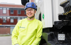 Nuori mies nojailee autoon ja nauraa. Takana näkyy punaisia rakennuksia. Miehellä on päässään sininen kypärä, suojalasit ja keltainen huomiopaita.