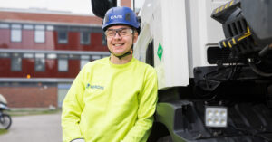 Nuori mies nojailee autoon ja nauraa. Takana näkyy punaisia rakennuksia. Miehellä on päässään sininen kypärä, suojalasit ja keltainen huomiopaita.