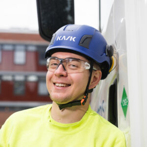 Sininen kypärä ja suojalasit päässä oleva mies hymyilee kameralle. Päällään hänellä on keltainen huomiopaita.