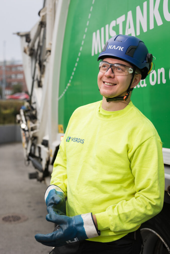 Sininen kypärä ja suojalasit päässä oleva mies hymyilee ja katsoo ylös kaukaisuuteen. Päällään hänellä on keltainen huomiopaita.