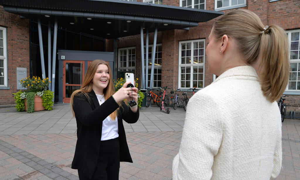 Nuori nainen pitää kädessään puhelinta ja kuvaa sillä toista naista. Takana näkyy punatiilistä rakennusta.