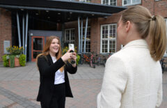 Nuori nainen pitää kädessään puhelinta ja kuvaa sillä toista naista. Takana näkyy punatiilistä rakennusta.