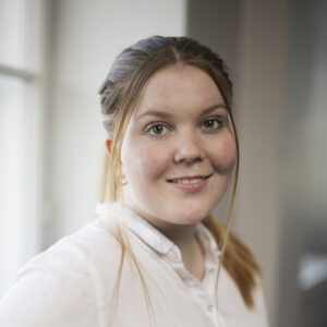 Nuori nainen hymyilee ja katsoo kameraan. Hänellä on päällään valkoinen rento kauluspaita.