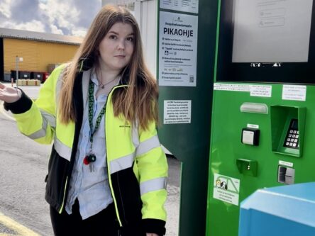 Nuori nainen seisoo vihreän automaatin vieressä hieman mietteliäänä käsi sivulle ojennettuna.