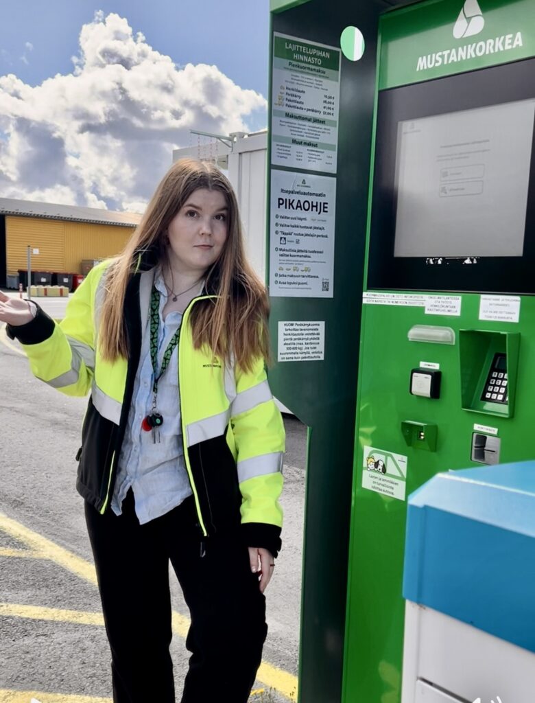 Nuori nainen seisoo vihreän automaatin vieressä hieman mietteliäänä käsi sivulle ojennettuna.