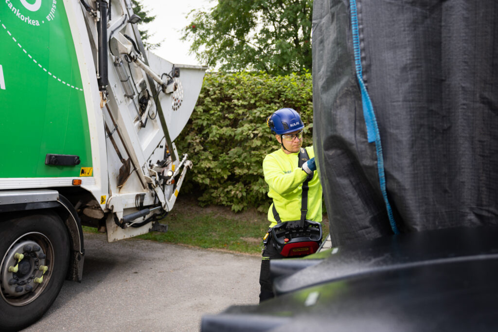 Etualalla näkyy mustaa isoa jätesäkkiä, jonka vieressä on huomiopaita päällä oleva kuljettaja. Taustalla näkyy vihreää jäteautoa ja viheraitaa.