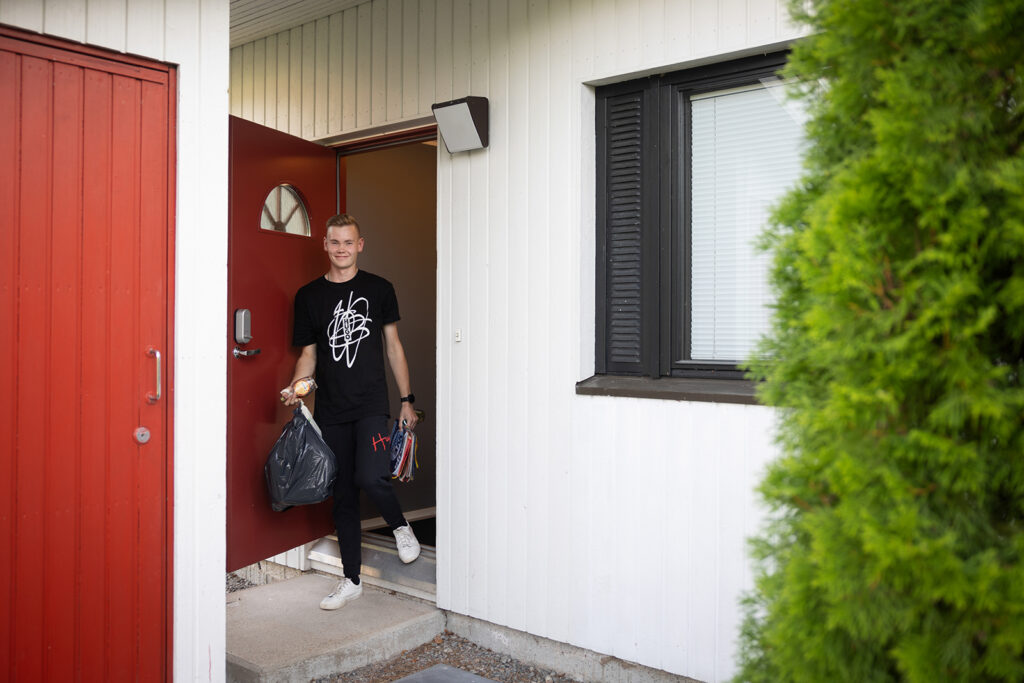 Nuori mies on tulossa rivitaloasunnosta ulos. Kädessä hänellä on roskapusseja. On kesä.