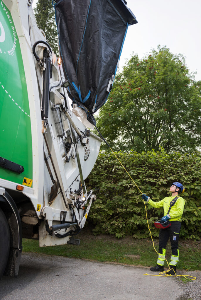 Ylhäällä näkyy iso musta jätesäkki, jota tyhjennetään jäteautoon. Nuori mies pitää kiinni keltaisesta narusta.