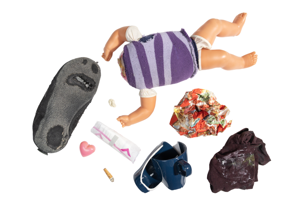 Kuvassa näkyy rikkinäinen nukke, lahjapaperia, rikkinäinen ja likainen vaate, rikkoutunut kahvikuppi, tupakantumppi, kuukautissuoja sekä rikkinäinen tossu.