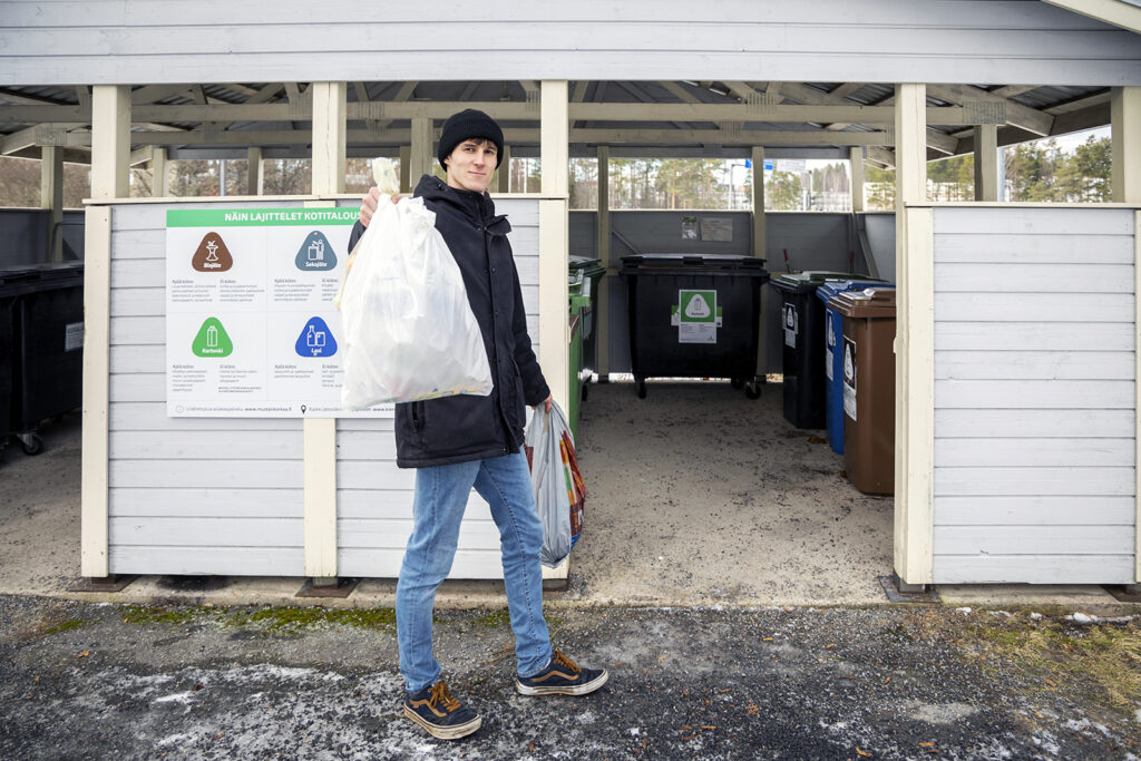 Nuori mies pitää ojentaa kädessään roskapussia. Takana näkyy taloyhtiön jätekatosta.