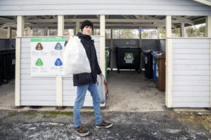 Nuori mies pitää ojentaa kädessään roskapussia. Takana näkyy taloyhtiön jätekatosta.
