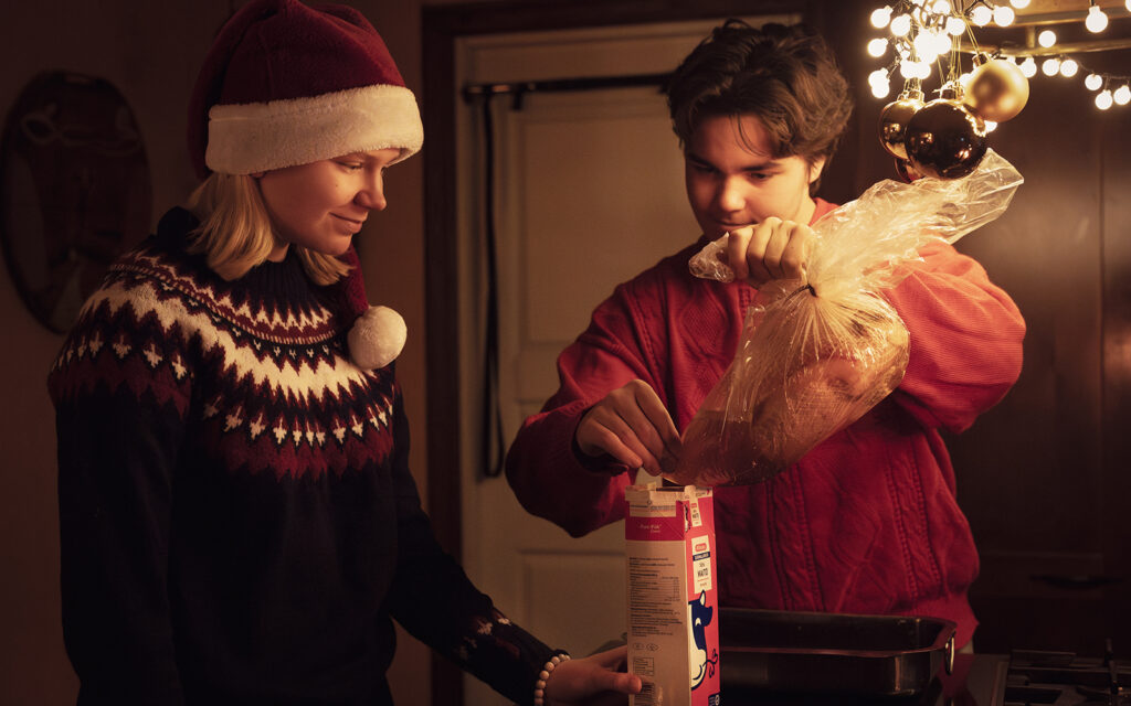 Tonttulakkipäinen nuori nainen pitää pöydällä punaista maitopurkkia. Punapaitainen nuorimies kaataa purkkiin jäähtynyttä kinkkurasvaa. On jouluinen tunnelma.