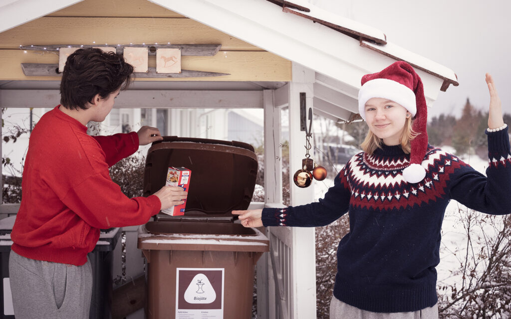 Tonttulakki päässä oleva nainen osoittaa biojäteastiaa, jonne punapaitainen nuo mies on tiputtamassa punaista maitotölkkiä.