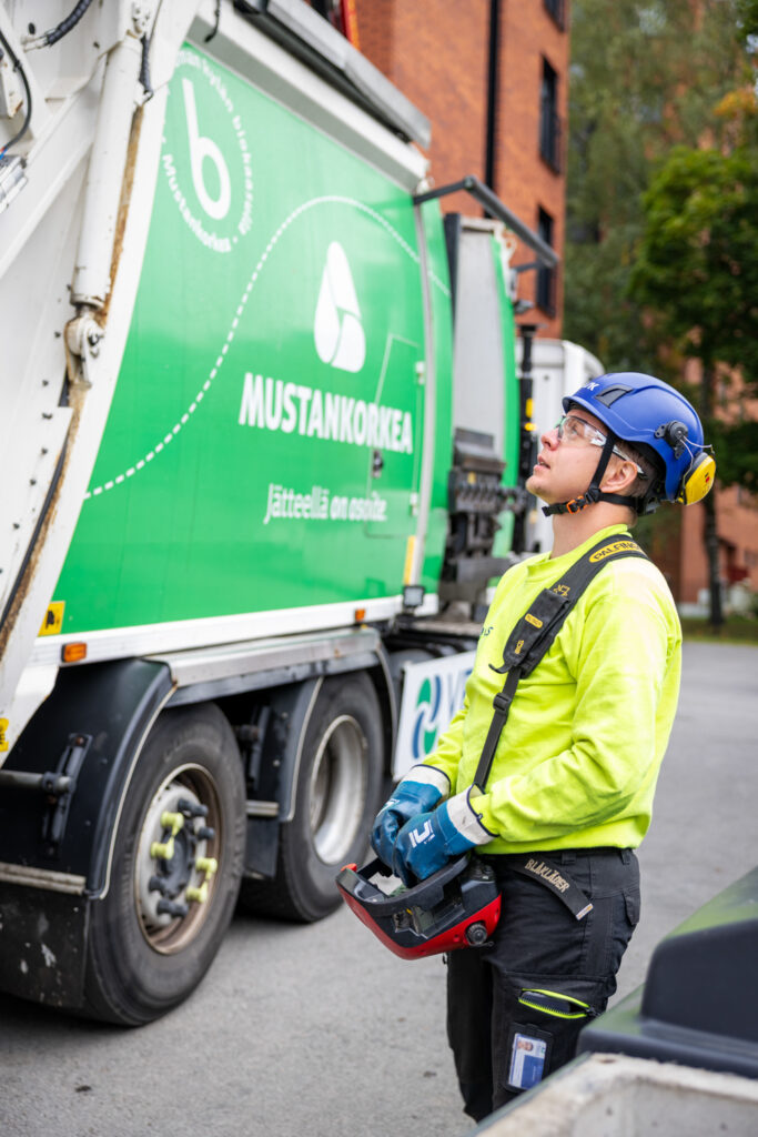 Nuori mies ohjaa kaukosäätimestä jotain ja katsoo ylöspäin. Takana näkyy vihreä jäteauto.