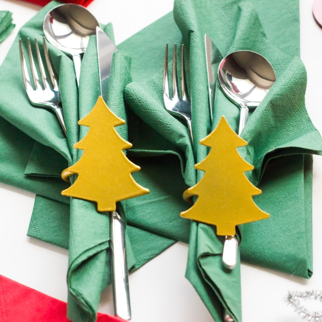 Pöydällä on vihreät servetit, joissa on lusikka, haarukka ja veitsi. Koristetaan on kultainen joulukuusi kattauksen päällä.