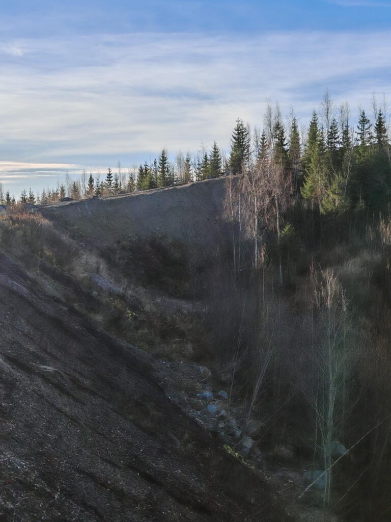 Kuvassa on todella jyrkkä penger, jonka takana näkyy puita ja alla hieman kiviä.