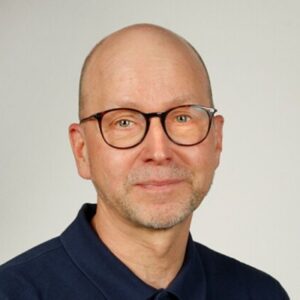 Silmälasipäinen mies katsoo kameraan. Päällään hänellä on tummapaita.