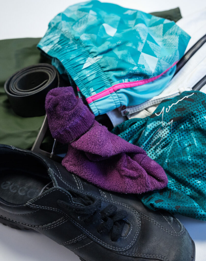 Kuvassa on musta kenkä, jotain urheiluvaatteita, vyö, violetti sukka ja taustalla valkoinen laukku.