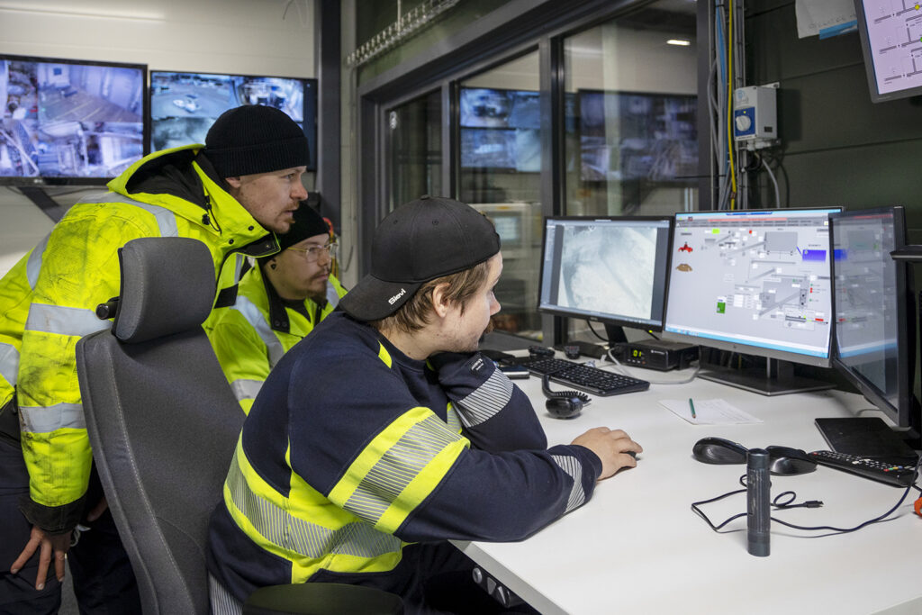 Kolme miestä katsoo kahta ruutua mietteliäänä. Myös taustalla näkyy lisää näyttöjä. Kaikilla on päällään jonkinlaiset huomiovaatteet.