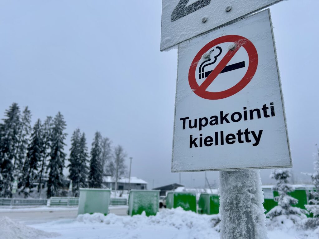 Kyltti, jossa lukee "Tupakointi kielletty". Taustalla näkyy vihreitä kontteja ja utuisia puita. Taivas on sinertävän harmaa.