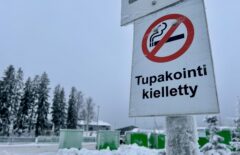 Kyltti, jossa lukee "Tupakointi kielletty". Taustalla näkyy vihreitä kontteja ja utuisia puita. Taivas on sinertävän harmaa.