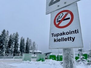 Kyltti, jossa lukee "Tupakointi kielletty". Taustalla näkyy vihreitä kontteja ja utuisia puita. Taivas on sinertävän harmaa.