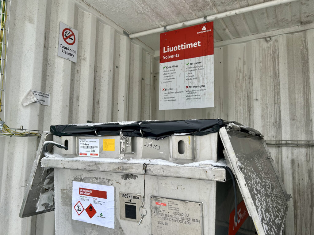 Valkoisessa rautakontissa on "Tupakointi kielletty" -kyltti, jonka vieressä on toinen isompi kyltti, jossa kerrotaan liuottimien lajitteluohjeet. Edessä näkyy rautakontti, jossa on vaarallisen jätteen huomiomerkkejä.