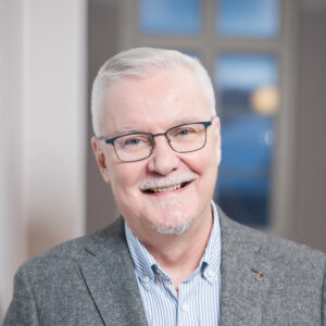 Silmälasipäinen mies katsoo hymyillen kameraan. Hänellä on päällään kauluspaita ja takki.