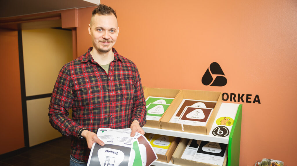 Ruutupaita päälle oleva mies pitää käsissään jäteastiatarroja. Taustalla näkyy niitä lisää hyllyssä.