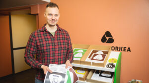 Ruutupaita päälle oleva mies pitää käsissään jäteastiatarroja. Taustalla näkyy niitä lisää hyllyssä.