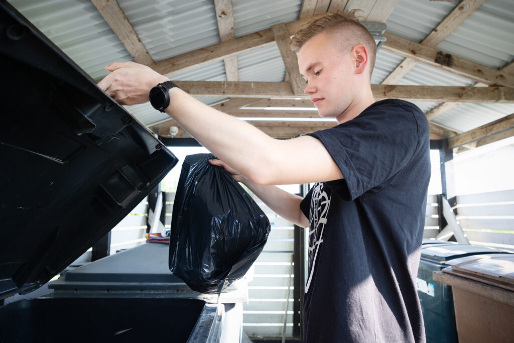 Nuori mies on t-paita päällä jätekatoksessa. Hänellä on kädessään musta pussi, jonka hän on viemässä roskikseen.