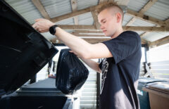 Nuori mies on t-paita päällä jätekatoksessa. Hänellä on kädessään musta pussi, jonka hän on viemässä roskikseen.